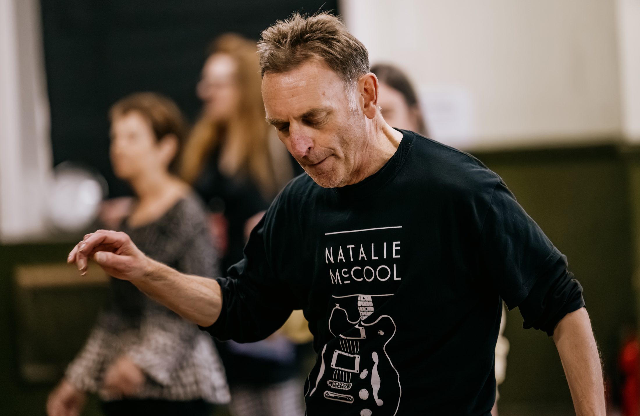 A man wearing a black Natalie McCool shirt with a guitar graphic is dancing indoors, with several blurred people in the background.