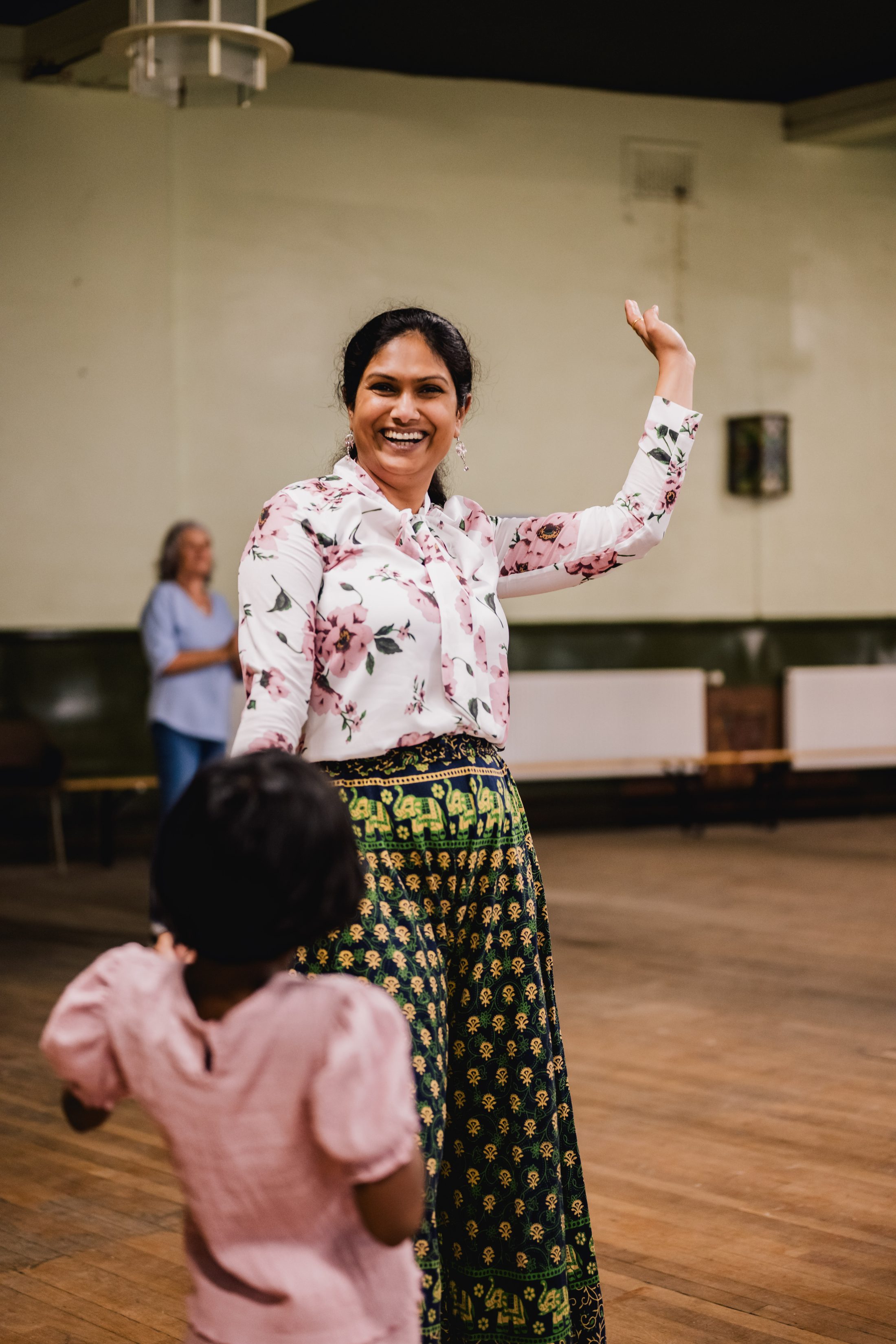 A smiling woman in a floral shirt and patterned skirt waves her arm joyfully whilst holding hands with a small child in a pink dress inside a large, empty room. Another woman stands blurred in the background.