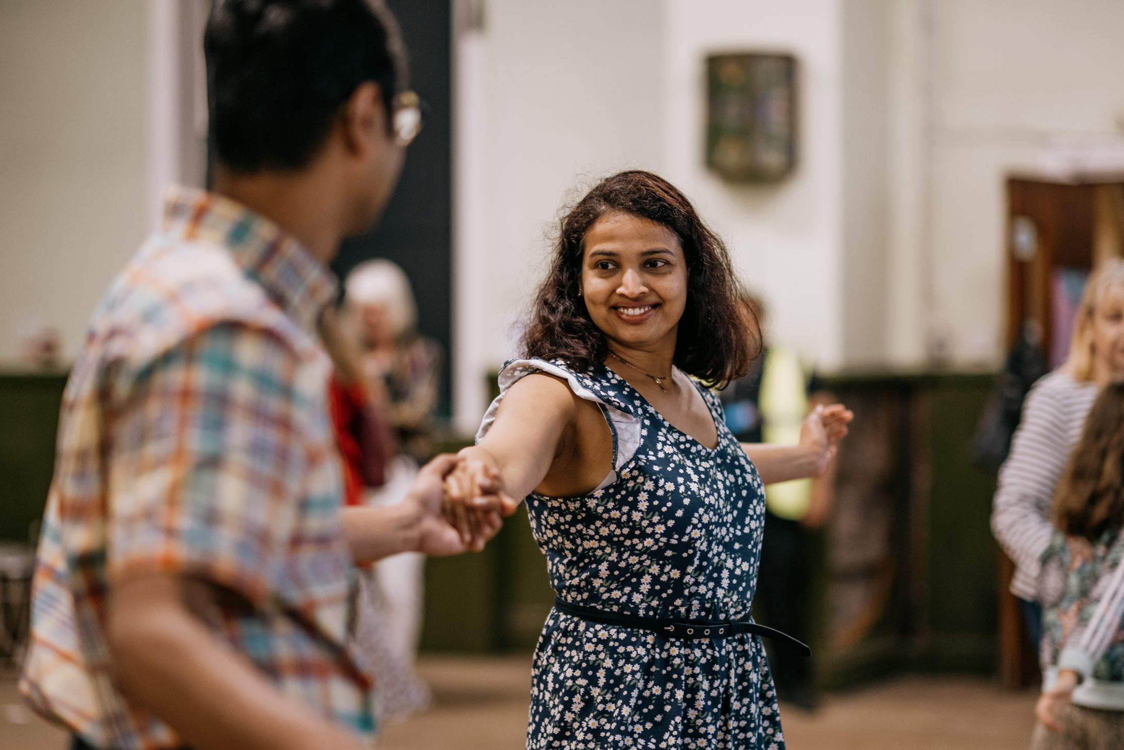 A woman in a blue floral dress smiles whilst dancing and holding hands with a man in a checked shirt at an indoor social event. Other people are visible in the background.
