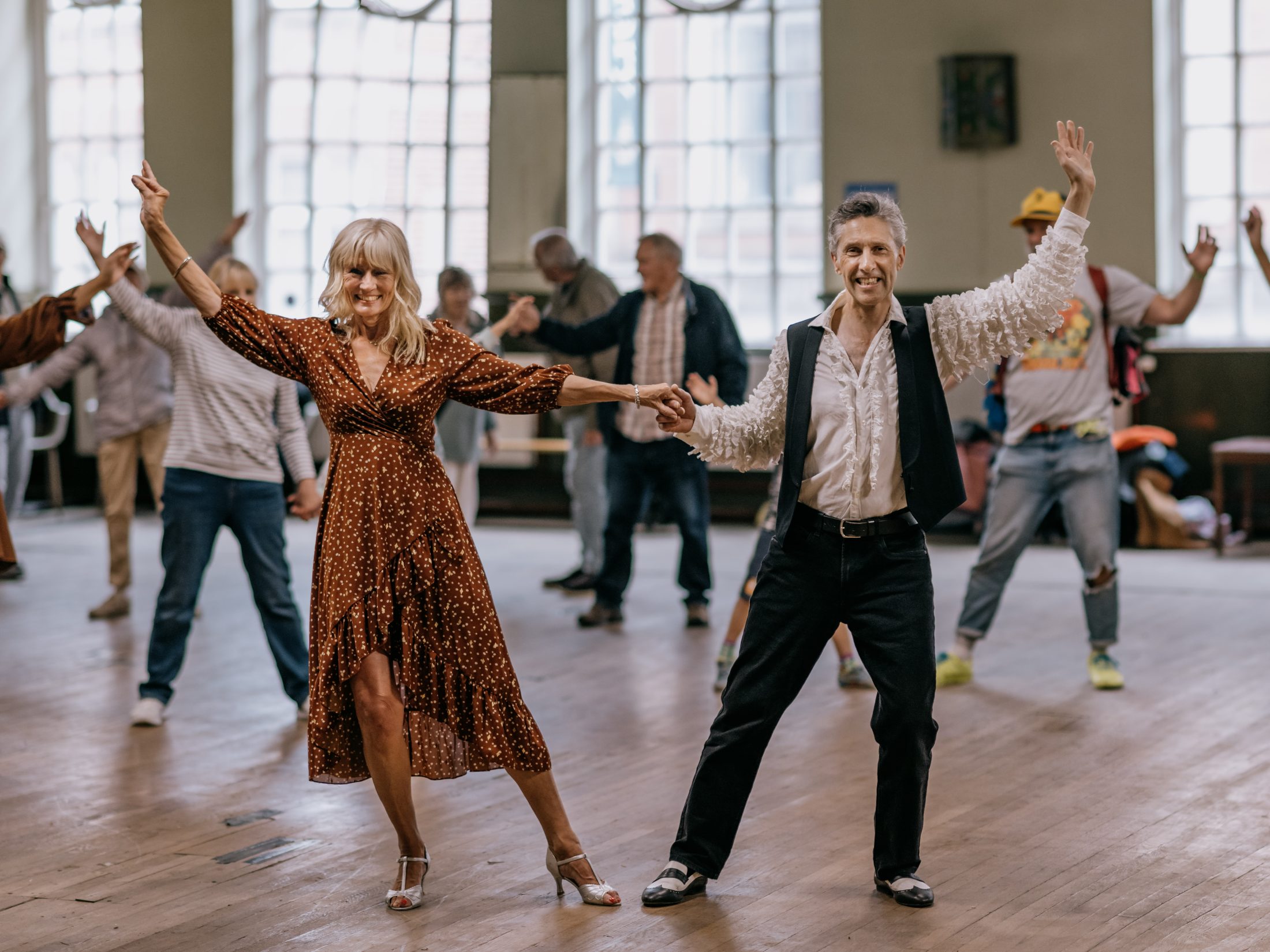 A group of people, led by a smiling older woman in a brown dress and a man in a waistcoat, dance in a bright, spacious room with large windows and wooden floors.