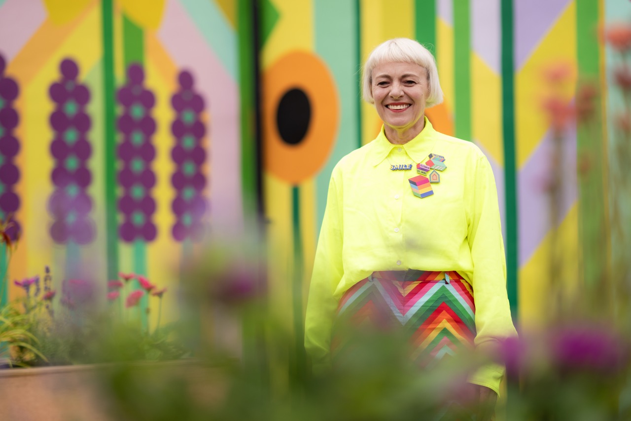 A smiling person with short blond hair wears a bright yellow shirt and a colourful, chevron-patterned skirt, standing in front of a vibrant, geometric mural with green, yellow, and purple shapes and flowers in the foreground.