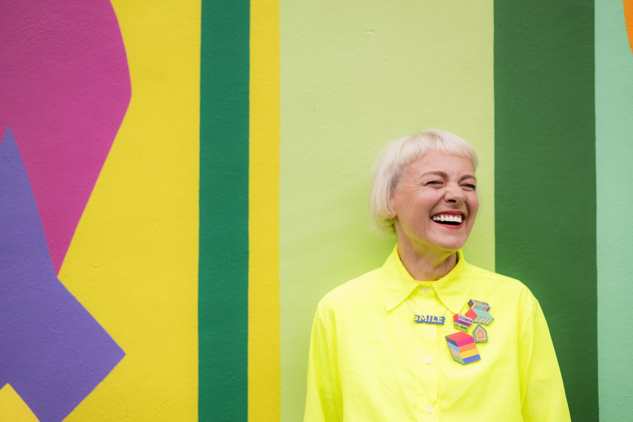 A joyful older woman with short white hair, wearing a bright yellow shirt with colourful accessories, smiles broadly whilst standing in front of a vibrant, multicoloured mural with geometric shapes.