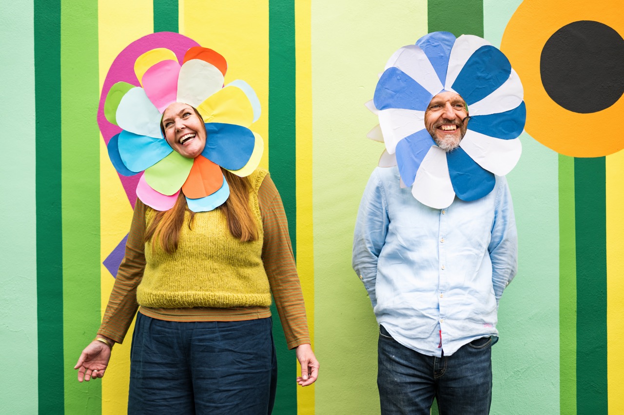Two smiling adults stand against a colourful mural, each wearing a large, playful flower costume around their heads—one with rainbow petals, the other blue and white. The background features green vertical stripes and bold flower designs.