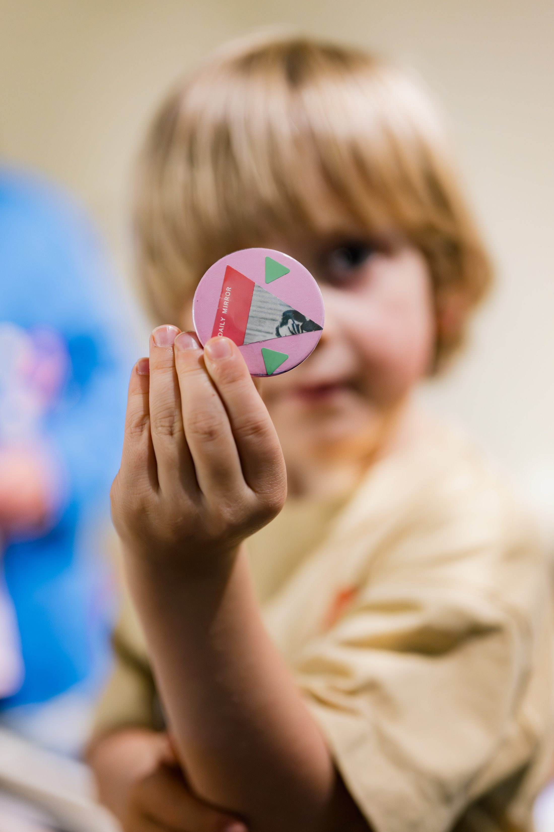 A young child with light brown hair holds up a round badge featuring the Statue of Liberty and pink and green colours, focusing the badge towards the camera while their face is slightly blurred in the background.
