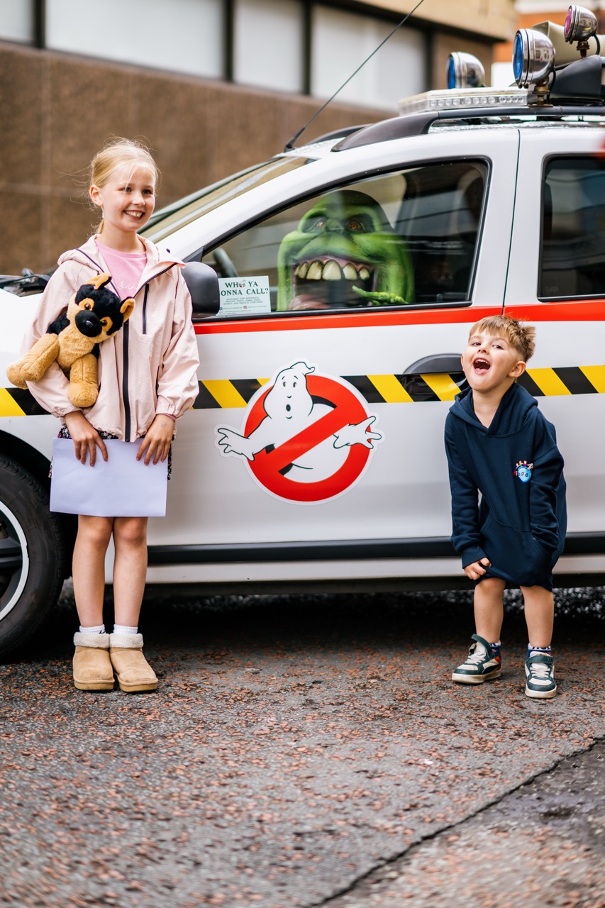 Two smiling children stand in front of a Ghostbusters car; the girl holds a stuffed Scooby-Doo toy, and a green ghost prop is in the driver's seat. The boy is laughing, and both look excited.