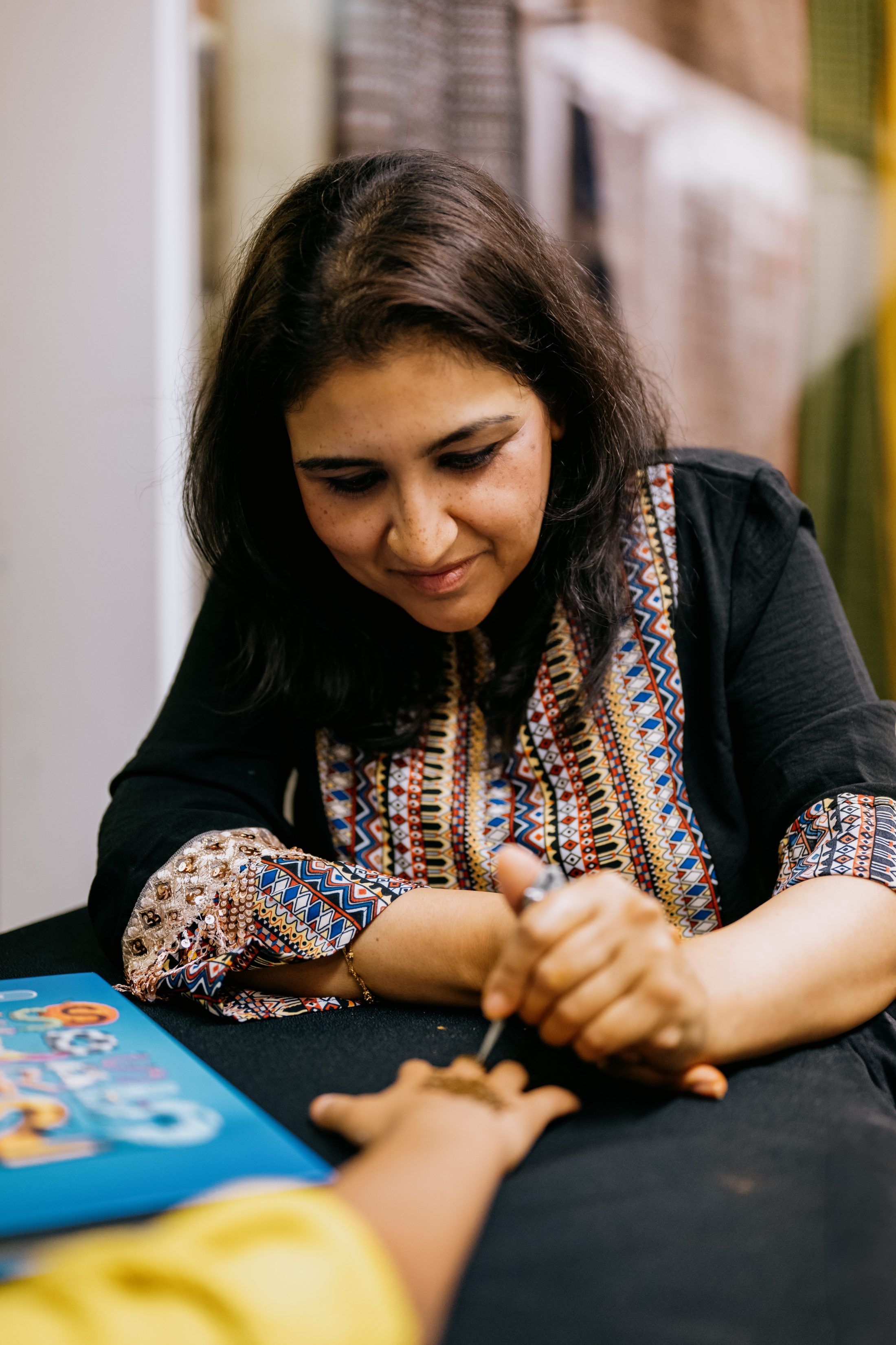 A woman in a patterned black top applies henna to someone's hand, focussing carefully on her work at a table with a colourful object nearby.