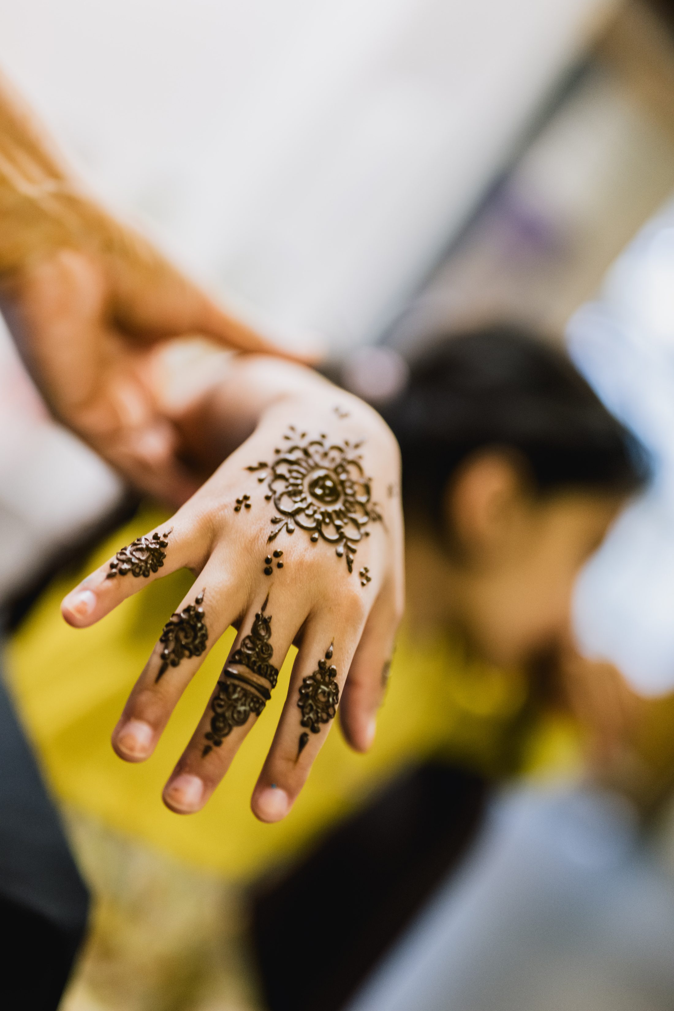 A close-up of a hand decorated with intricate mehndi (henna) designs, featuring floral and geometric patterns, with a blurred figure in the background.