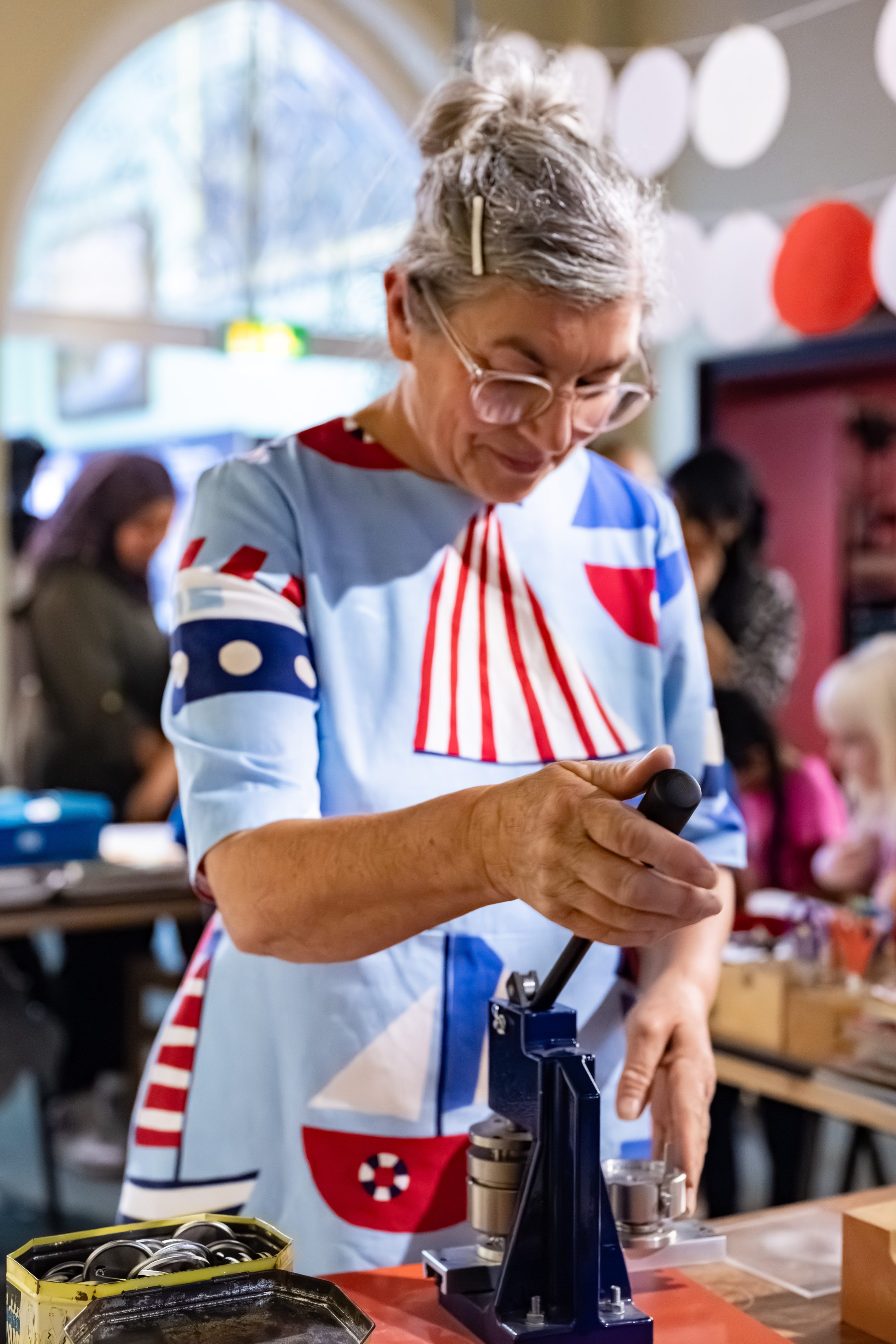 A woman with grey hair and glasses, wearing a blue, red, and white nautical-print dress, operates a badge-making press at a busy indoor craft event.