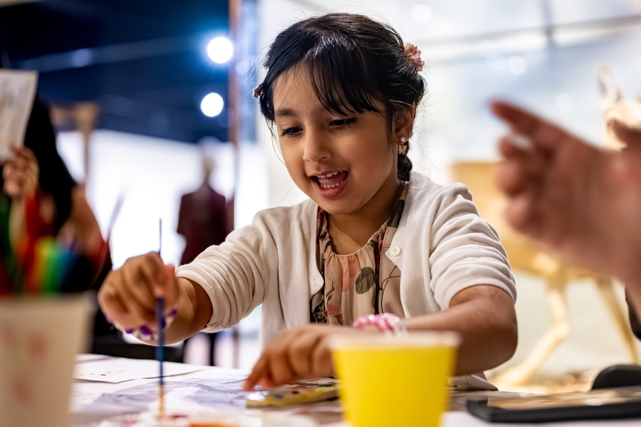 A young girl smiles whilst painting at a table. She holds a paintbrush and reaches towards art supplies. The background is softly blurred, suggesting a creative indoor setting.