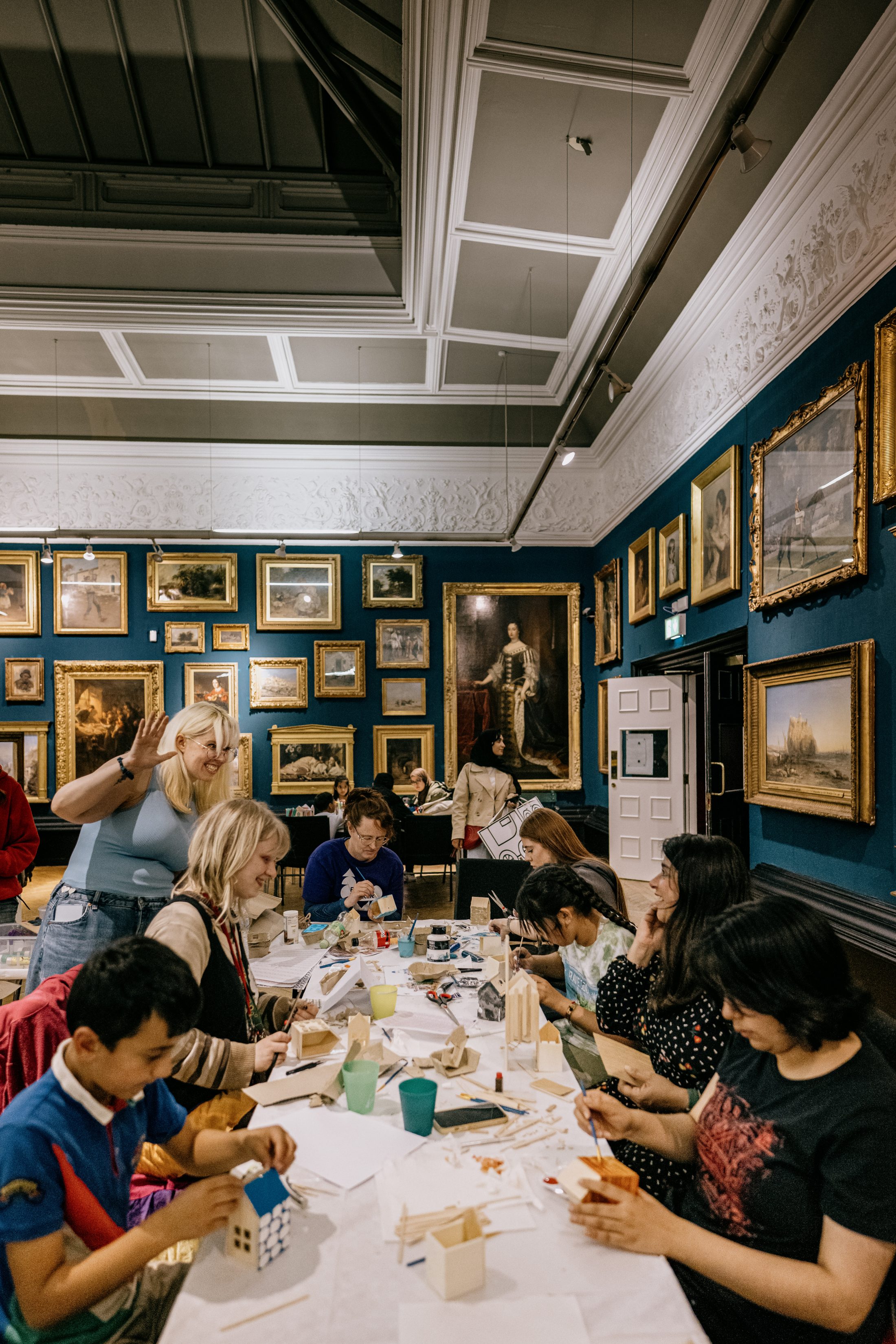 A group of adults and children sit around tables in an art gallery, working on crafts. Framed paintings cover the walls, and materials like paper, wood, and paint are scattered on the tables.