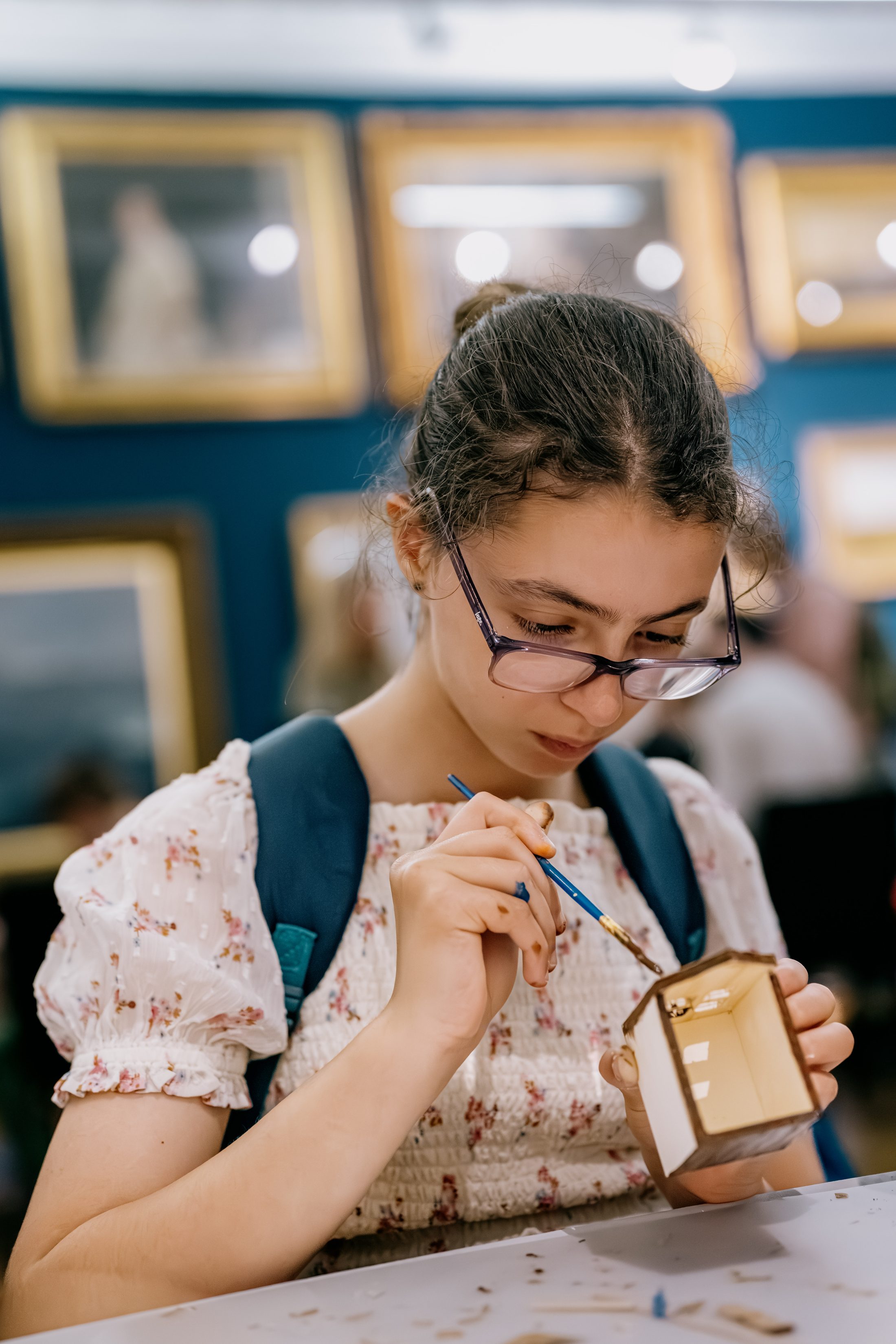 A young person with glasses and a blue rucksack focuses on painting a small wooden house, sitting at a table in a room decorated with framed artwork.