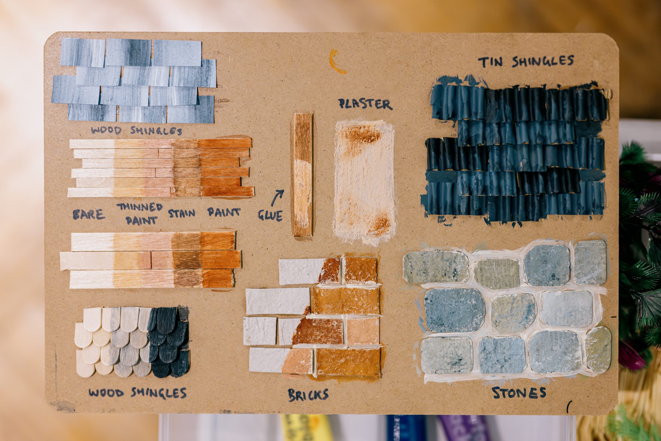 A board displays labelled samples of miniature building materials: wood shingles, tin shingles, painted wood pieces, plaster, bricks, and stones, each showing different textures, colours, and finishes for reference.