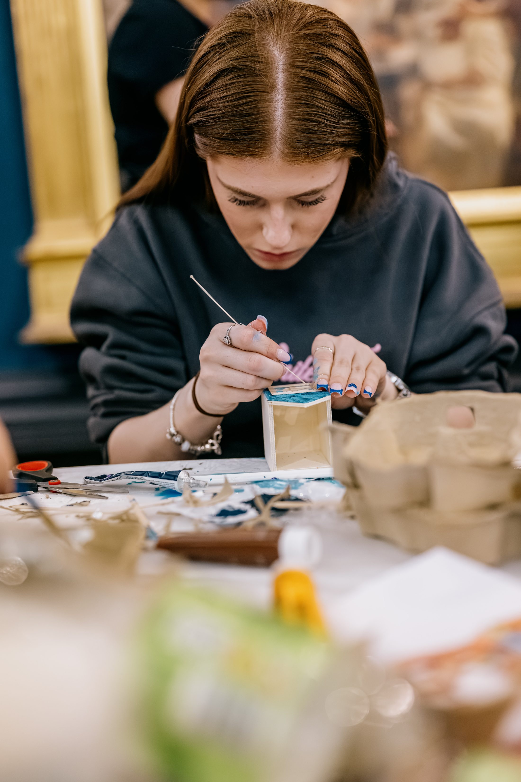 A young woman with brown hair focuses intently as she paints a small wooden box at a table covered with art supplies and craft materials.