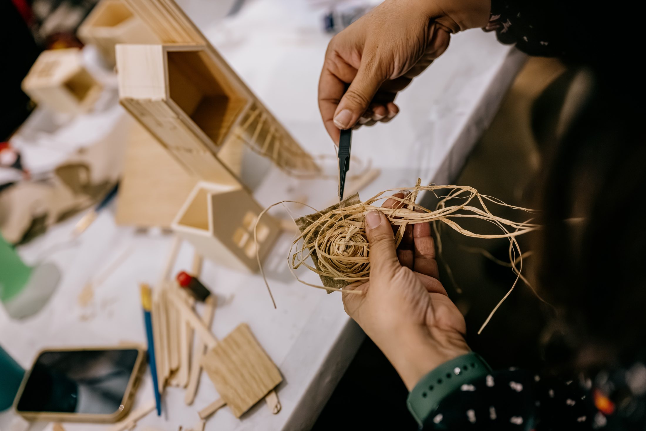 Person’s hands crafting with wood pieces and straw at a table, using a small tool. Various wooden parts and crafting materials are spread around the workspace.