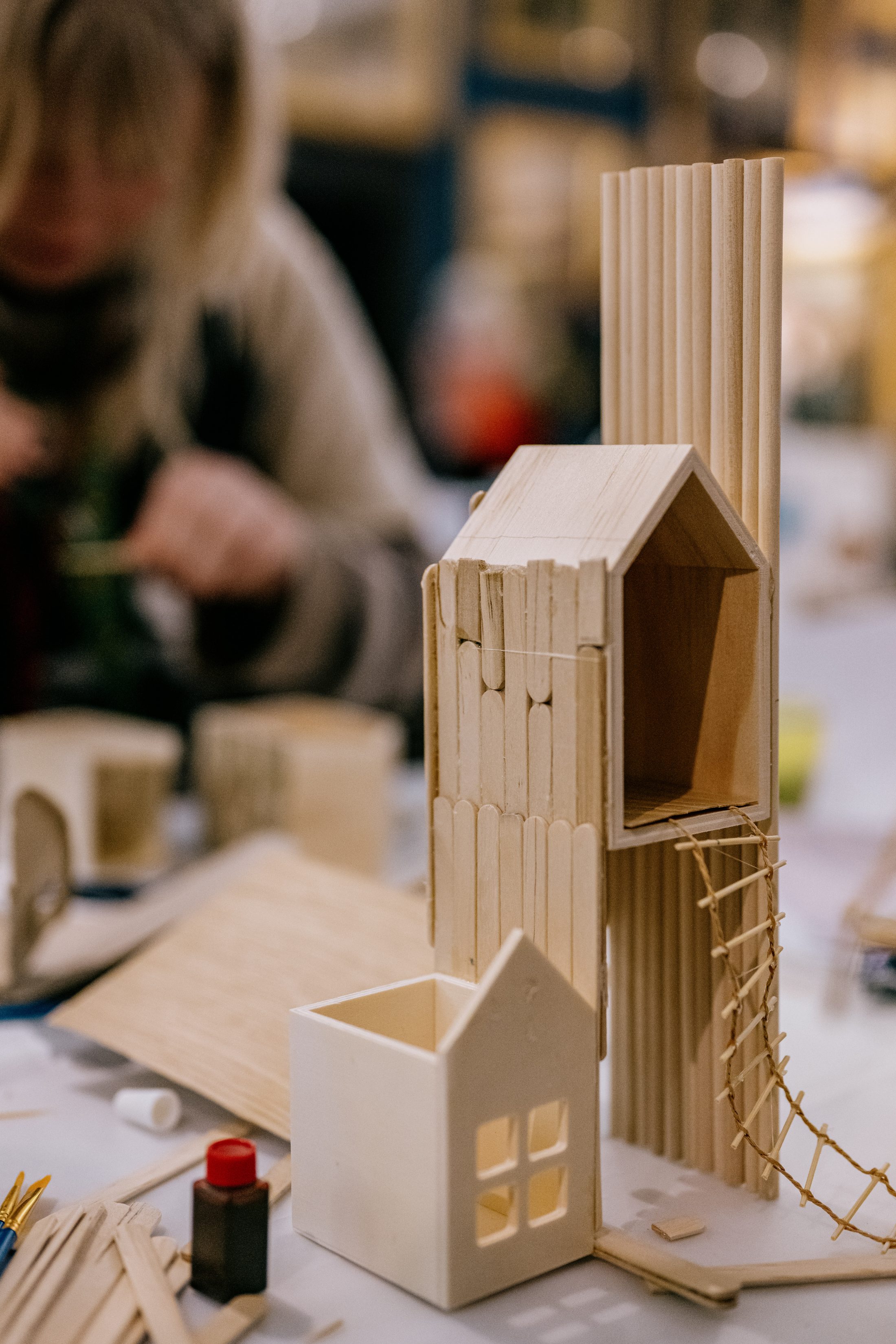 A close-up of a miniature wooden house model with sticks and panels, featuring a rope ladder. In the blurred background, a person is assembling another model on a table. Craft supplies are scattered around.