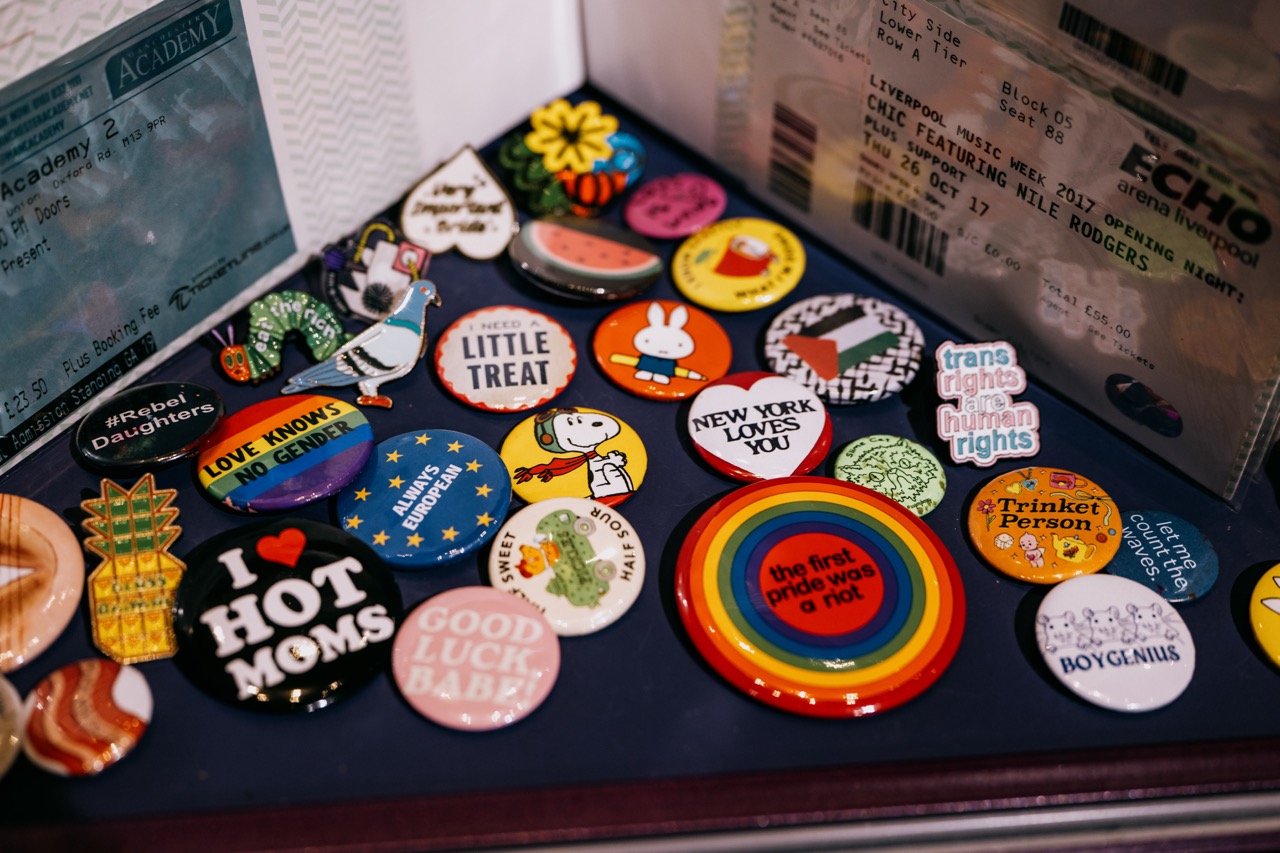 A collection of colourful pins and badges with various slogans and images, such as rainbows, cartoon characters, and positive messages, arranged next to concert tickets and other memorabilia.