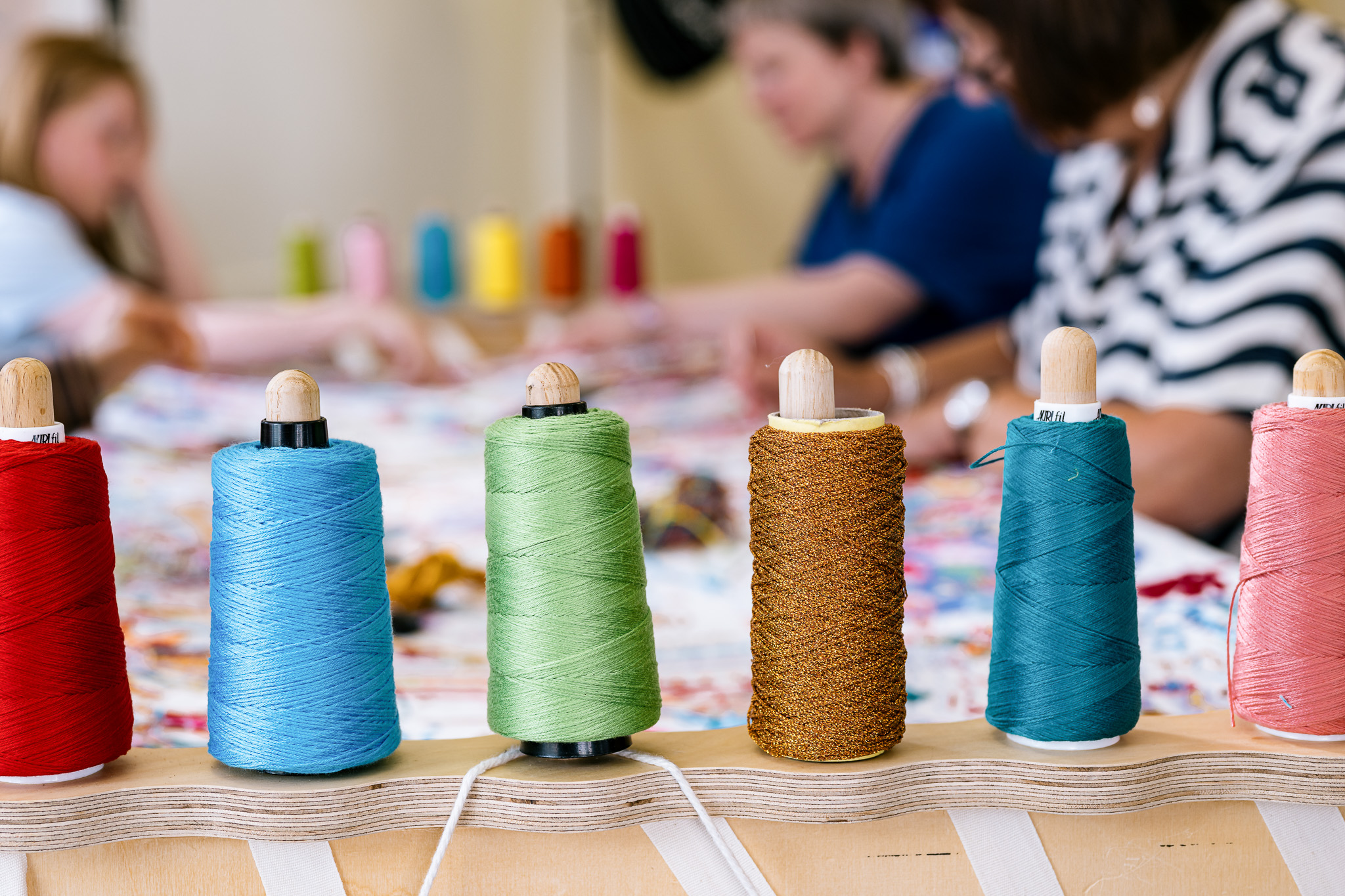 Spools of colorful thread in red, blue, green, gold, and teal are lined on a wooden holder. In the blurred background, people are engaged in stitching.