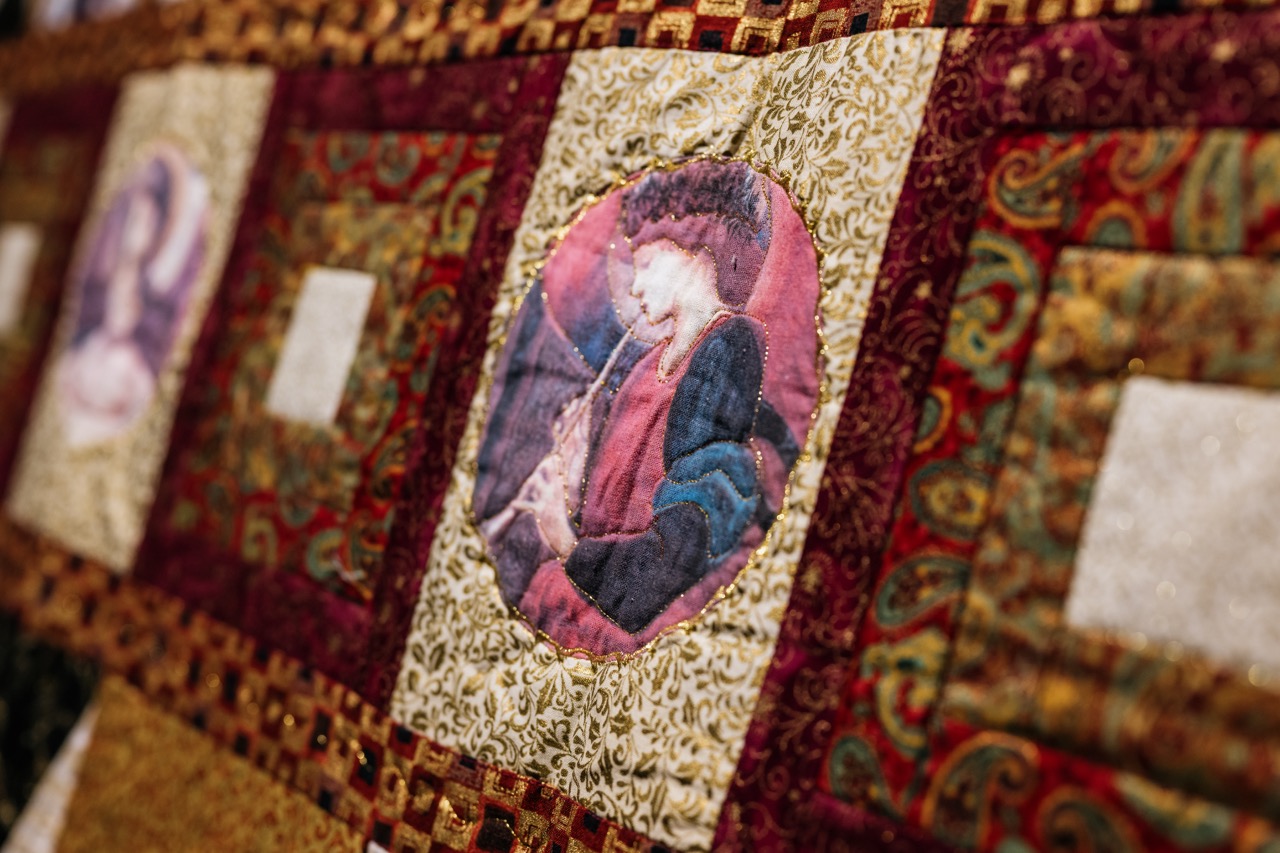 A close-up of a quilt featuring a patchwork design with ornate patterns and a central fabric portrait of a person in historical clothing, surrounded by red, gold, and green fabric squares.