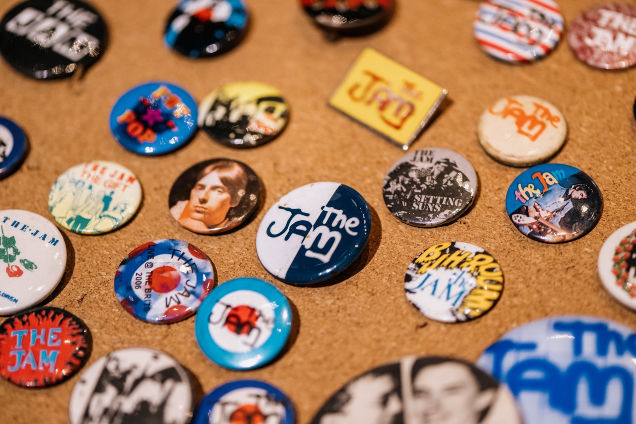 A collection of colourful band badges and buttons featuring The Jam and various designs and images, displayed on a corkboard background.