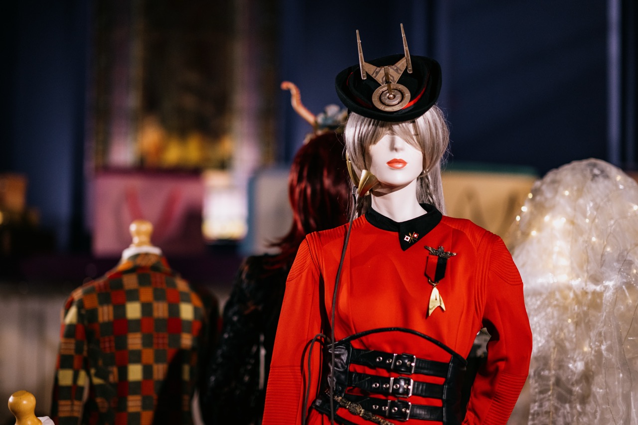 A mannequin dressed in a futuristic red outfit with a Star Trek badge, black belts, and an elaborate headpiece stands in focus, with other mannequins and blurred, colourful background elements behind it.