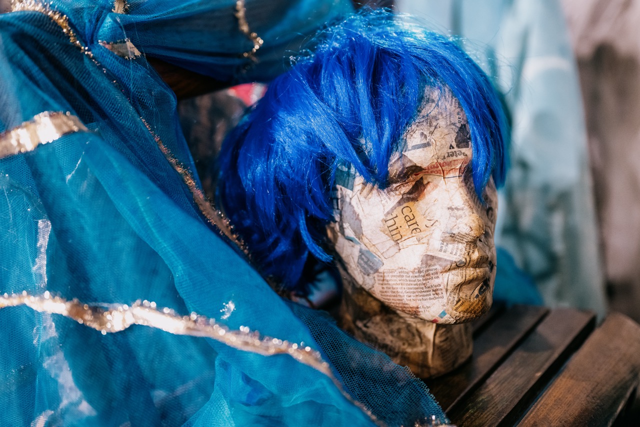 A mannequin head covered in newspaper cuttings wears a bright blue wig, surrounded by blue fabric with silver trim, resting on a wooden surface.