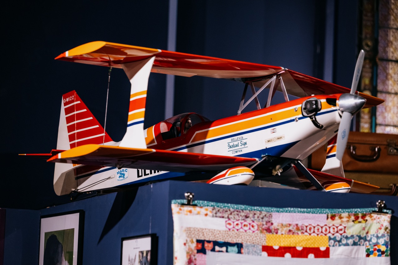 A brightly coloured model biplane with red, orange, and white stripes is displayed indoors on a blue shelf above framed pictures and a patchwork quilt.