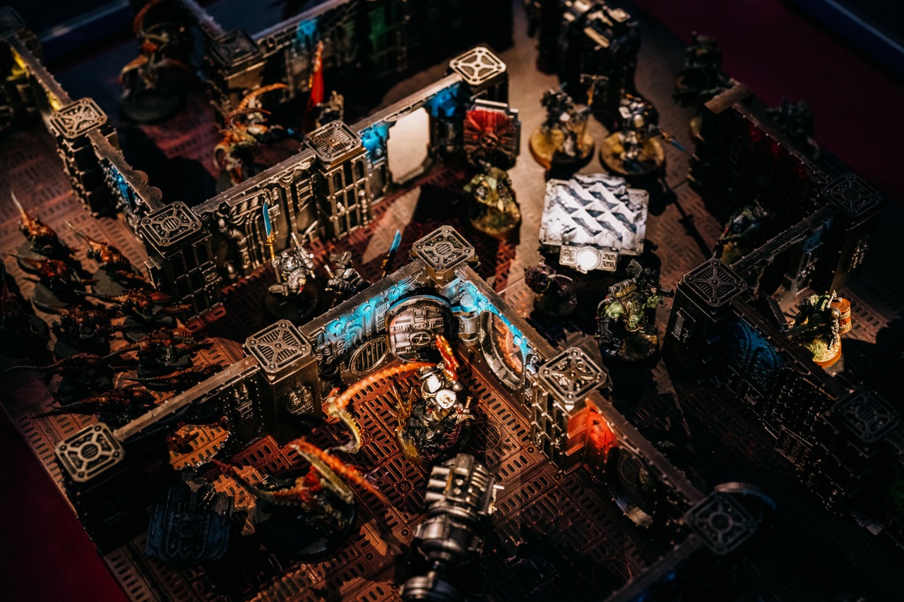 Tabletop wargame setup with detailed miniature figures and terrain pieces, showing a battle scene with walls, sci-fi structures, and painted figures engaged in combat across the board.