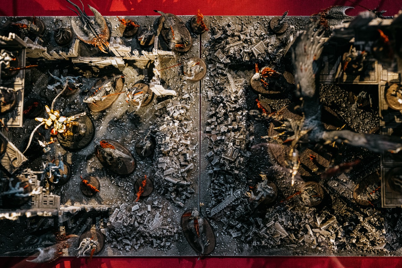 Tabletop miniature war game scene viewed from above, featuring detailed painted fantasy figures, ruins, and rubble arranged on a divided, textured terrain board with a red background.