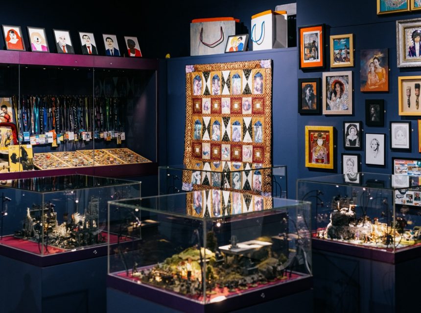 A museum exhibition with framed portraits, colourful quilts, display cases featuring miniature dioramas, and a wall with medals and lanyards, all arranged in a brightly lit room with dark blue walls.