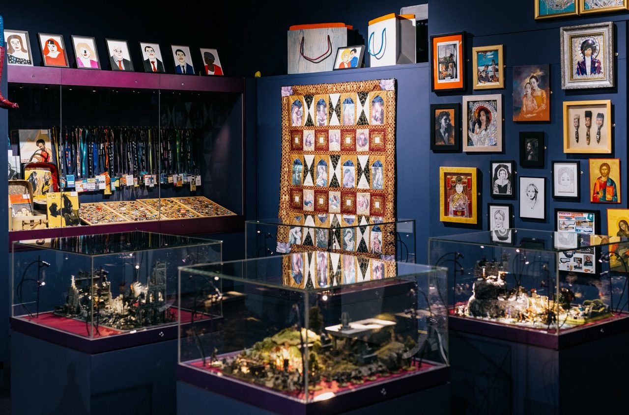 A museum exhibition with framed portraits, colourful quilts, display cases featuring miniature dioramas, and a wall with medals and lanyards, all arranged in a brightly lit room with dark blue walls.
