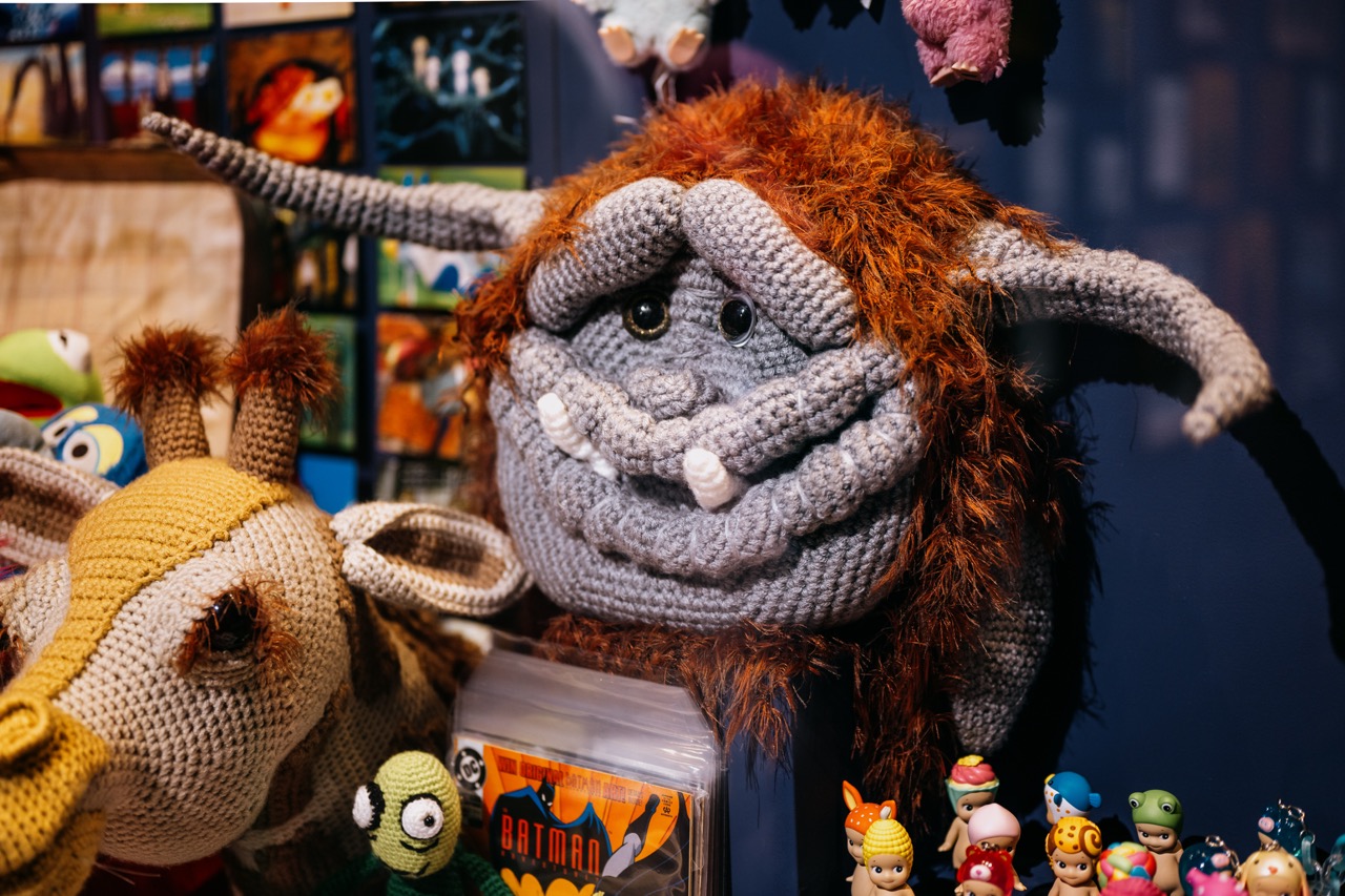 A display of crocheted toys, including a large grey creature with tusks and orange hair, a crocheted giraffe, and various collectable figurines on a shelf. Comic books are partially visible below the toys.