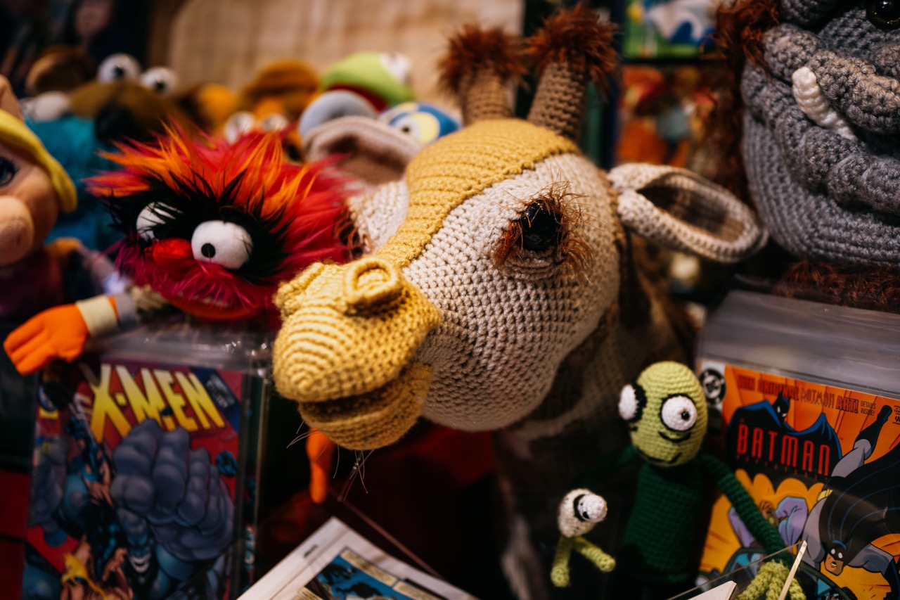 A close-up of a crocheted giraffe surrounded by various colourful handmade puppets and stuffed toys, with comic books featuring X-Men and Batman visible in the background.