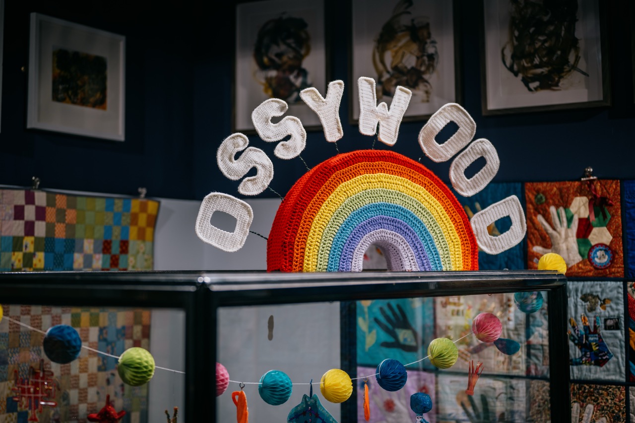 A colourful crocheted rainbow arch with large white letters spelling OSSYWOOD is displayed above a glass case. Multicoloured balls and quilts appear in the background, along with framed artwork on the wall.