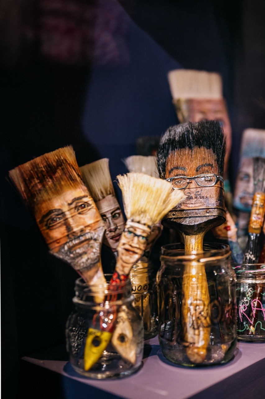 Paintbrushes with bristles painted as human faces are displayed upright in glass jars, creating a whimsical and artistic arrangement. The background is dark, making the colourful faces stand out.