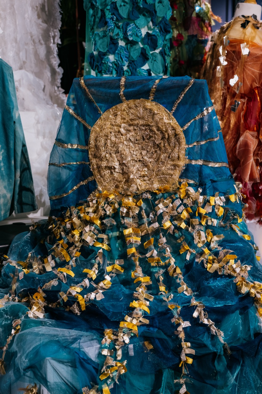 A blue fabric backdrop features a textured gold sun at the centre with gold and silver ribbons radiating outward, resembling sunrays, surrounded by colourful costumes and decorations in the background.