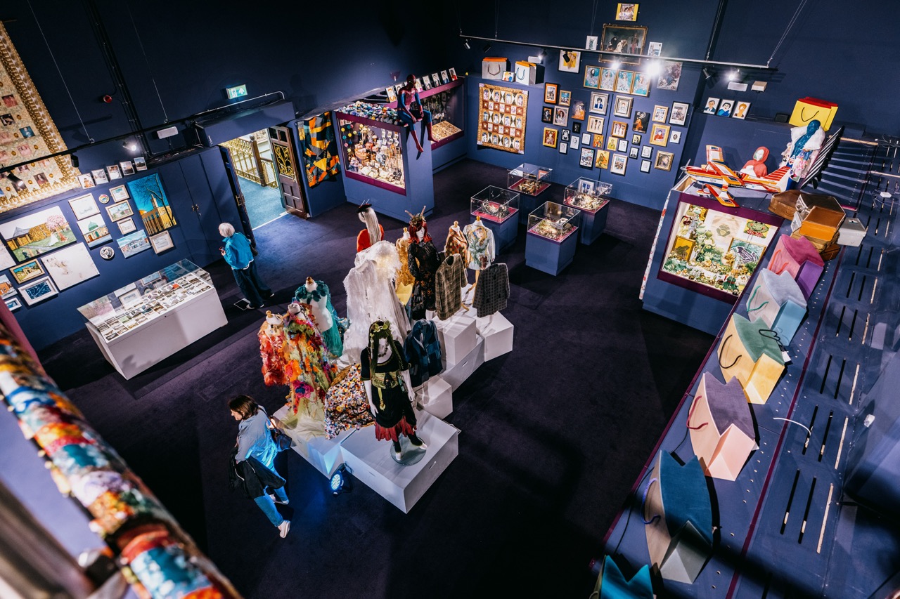 A museum gallery displays colourful costumes on mannequins, framed artworks on deep blue walls, glass cases with artefacts, and visitors exploring the exhibits. The space is well-lit and creatively arranged.