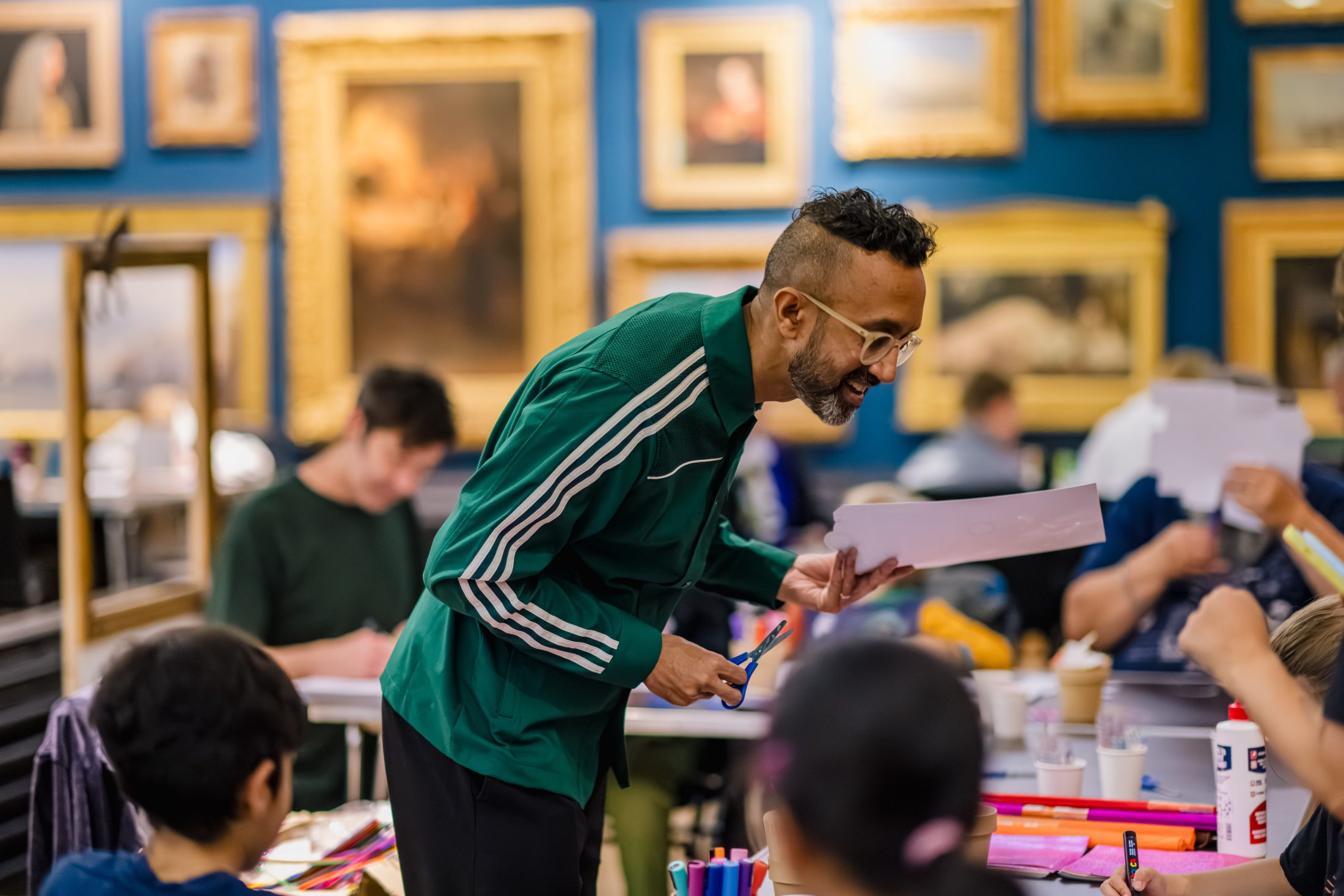 A smiling person in a green jacket interacts with children at a table covered in art supplies, with framed paintings hanging on the wall behind them in a classroom or museum setting.