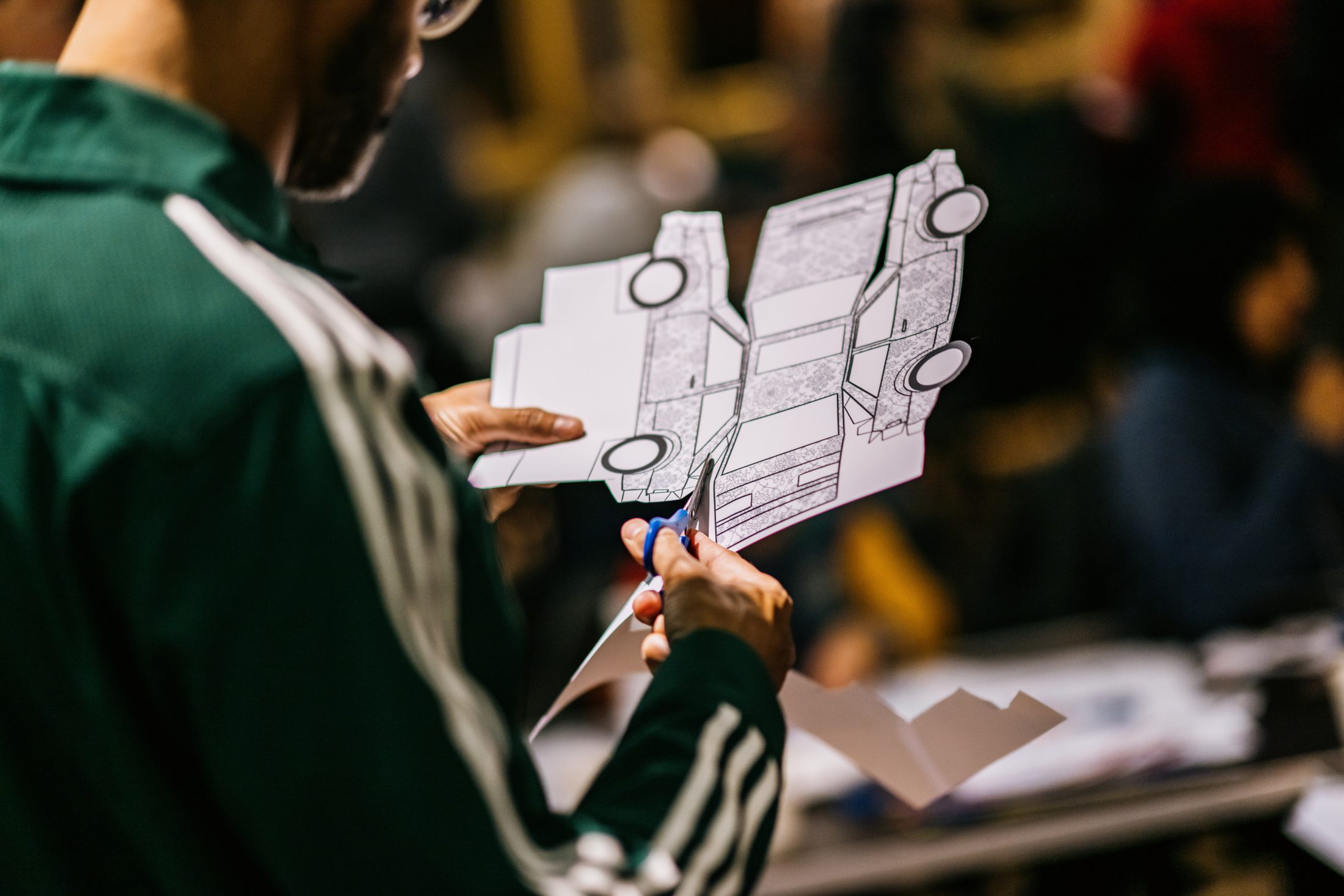 A person wearing a green jacket uses scissors to cut out a printed paper template of a car, preparing to assemble it into a model. The background is blurred with other people and tables visible.