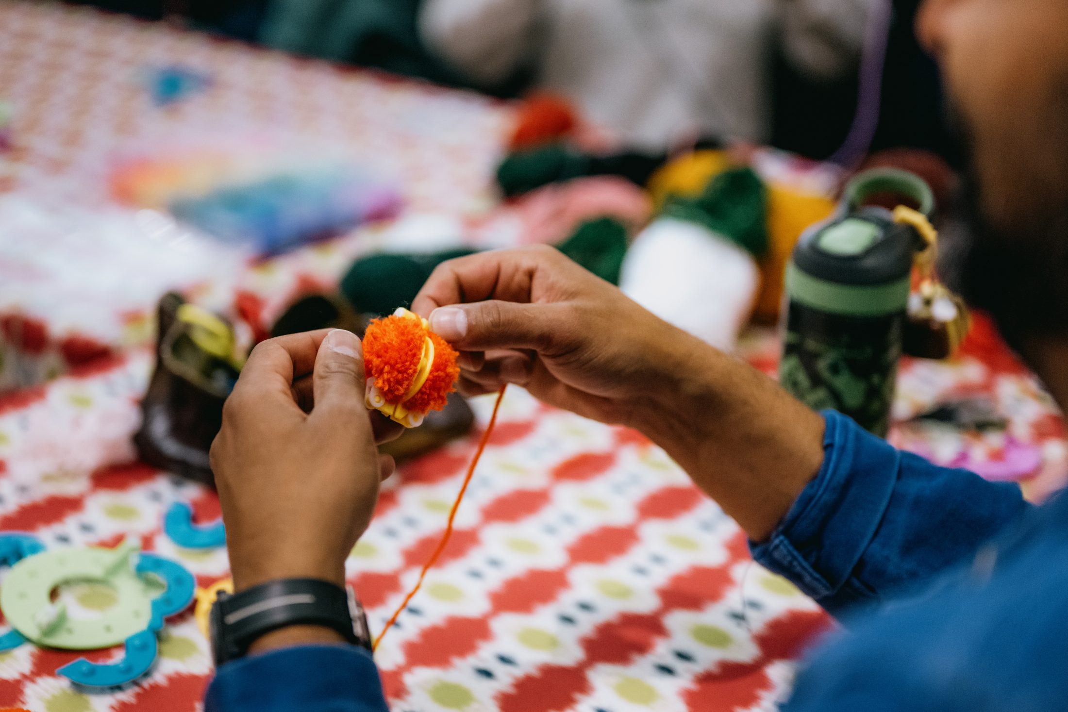 A person holds and crafts a red and yellow yarn pompom at a table covered with a colourful patterned cloth, surrounded by yarn, crafting tools, and a green water bottle.