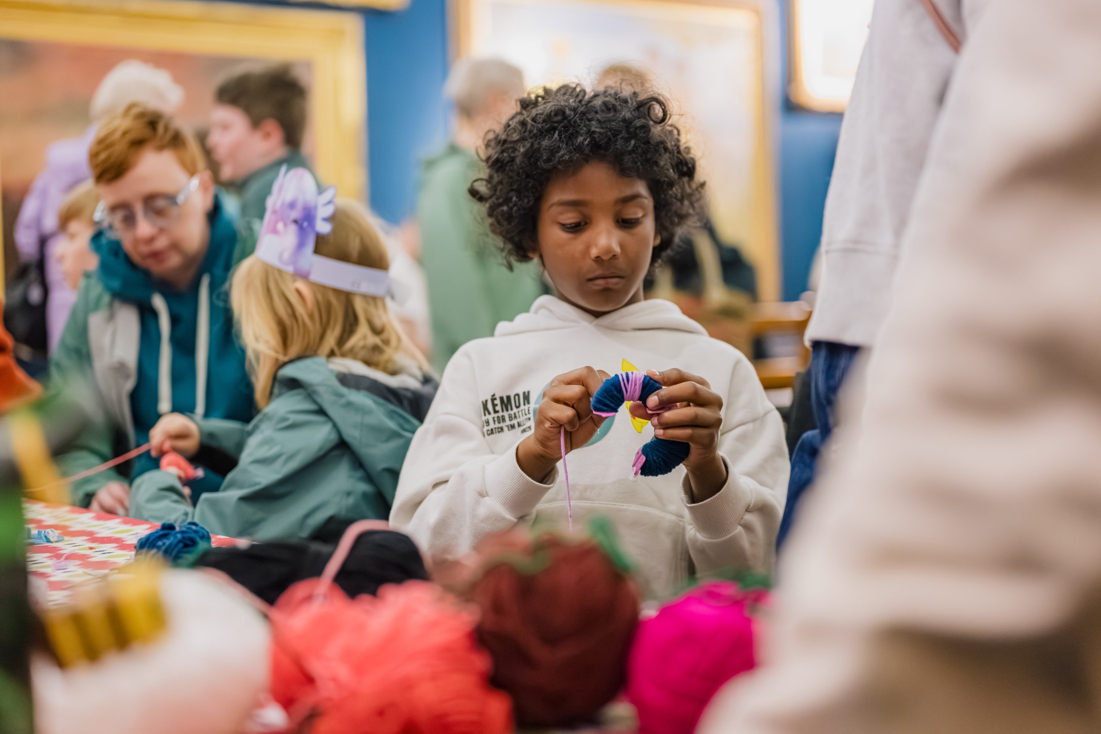 A child with curly hair concentrates on crafting with colourful yarn at a busy indoor event, whilst other children and adults work on projects around a table in the background.