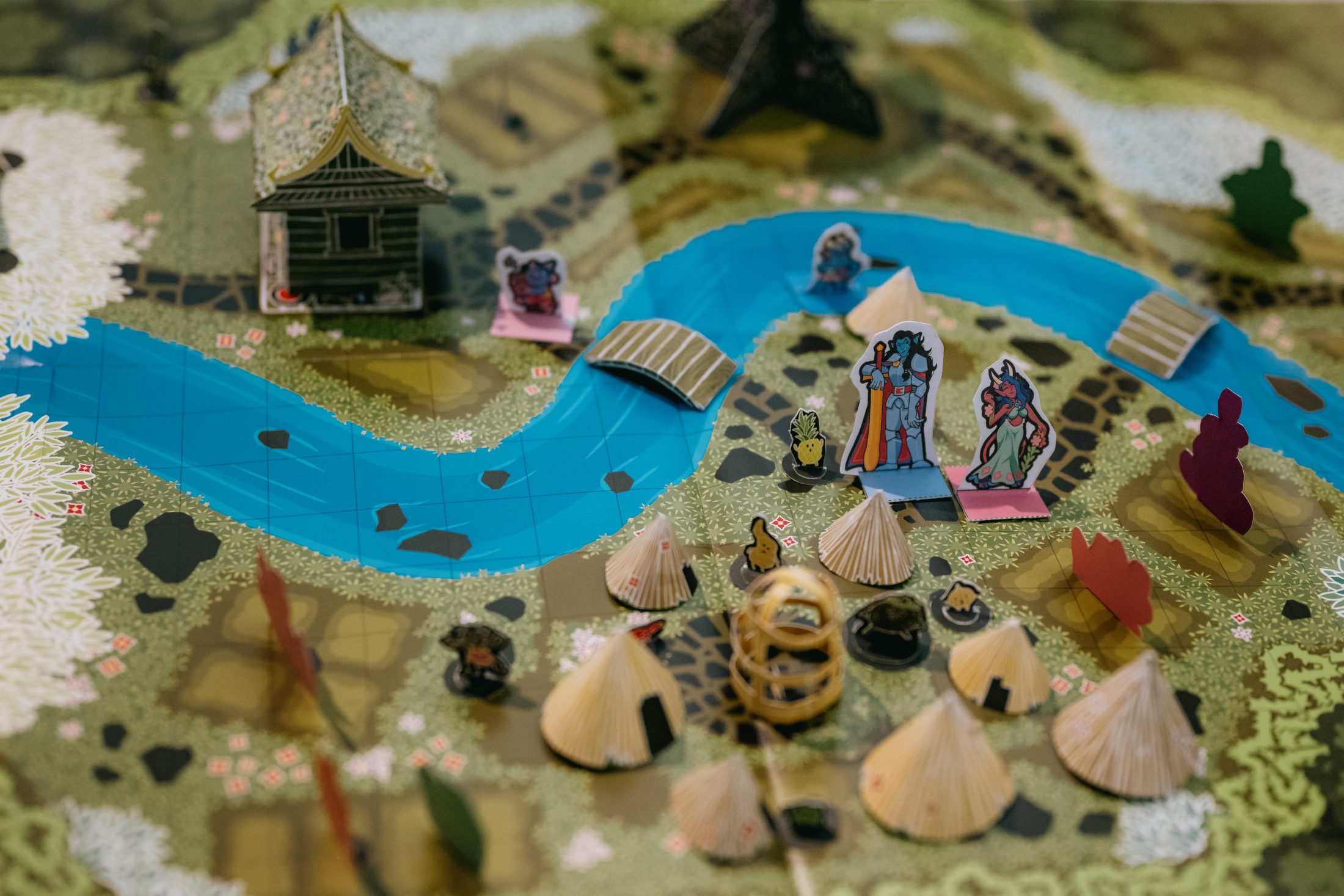 A colourful board game with a river, bridges, a small pagoda, and paper tents. Cardboard character standees and game pieces are arranged around the board on a detailed landscape map.