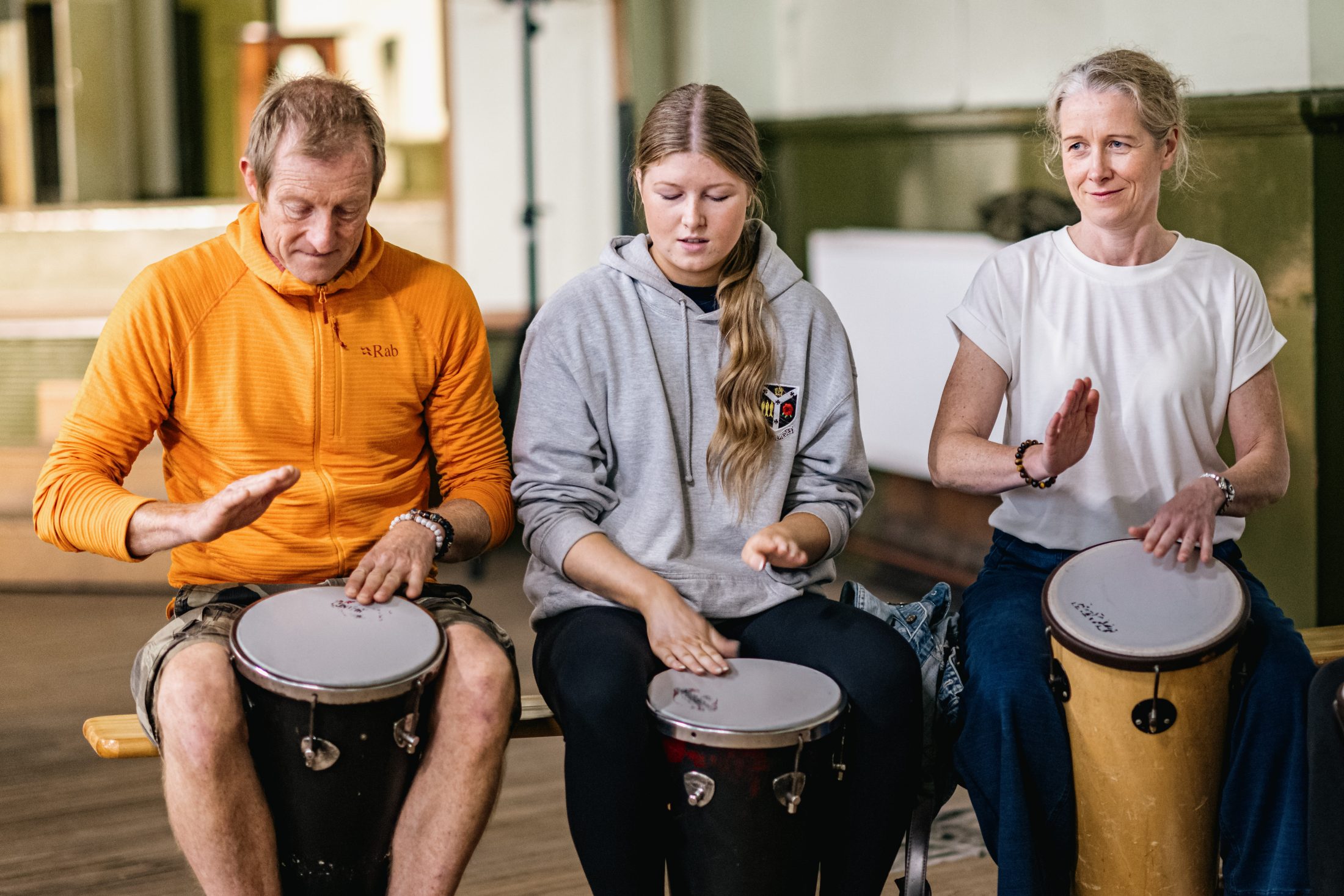 Three people sit in a row playing hand drums. The person on the left wears an orange hoodie, the person in the middle wears a grey hoodie, and the person on the right wears a white T-shirt. They all look focused on drumming.
