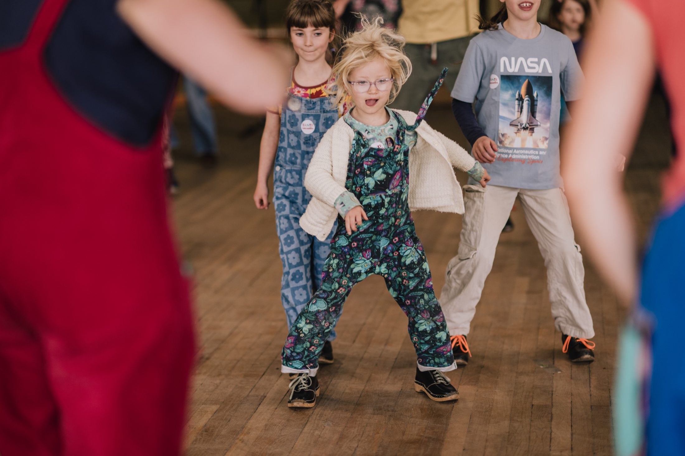 Children energetically dancing together indoors on a wooden floor, with one child in front wearing patterned dungarees and glasses. Other kids and adults are in the background, some partially out of frame.