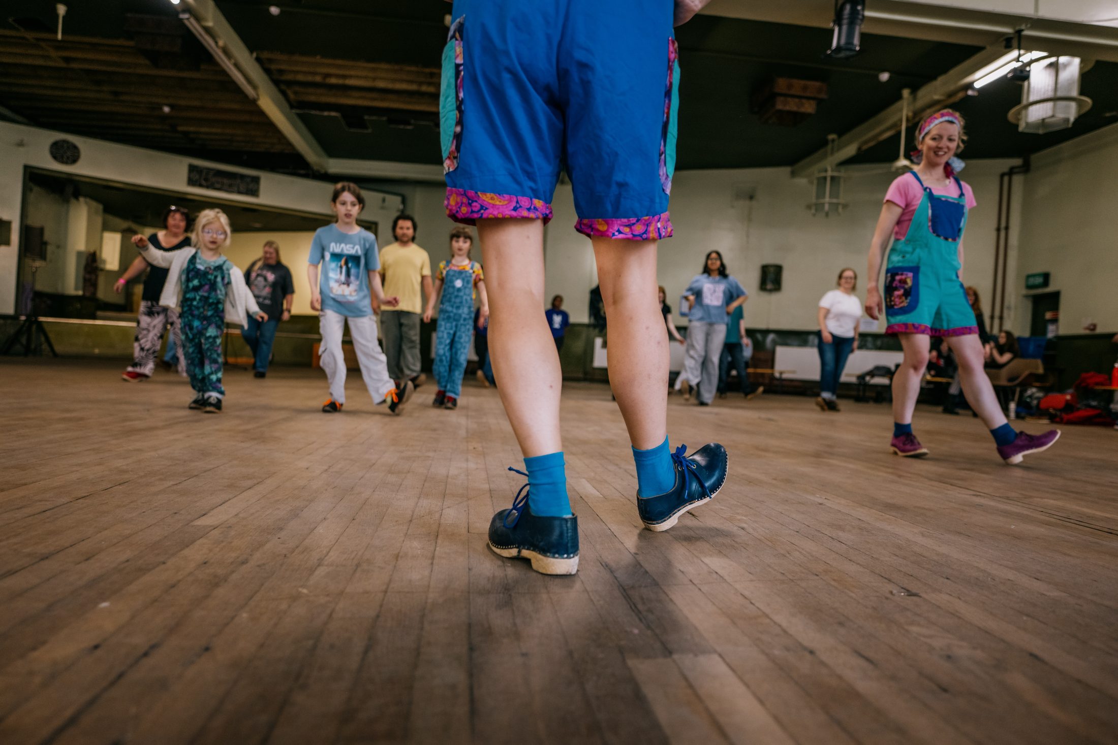 A group of people, including children and adults, are practising dance steps in a spacious hall with a wooden floor. The focus is on the legs of a dancer in blue shoes at the front, with others following in the background.