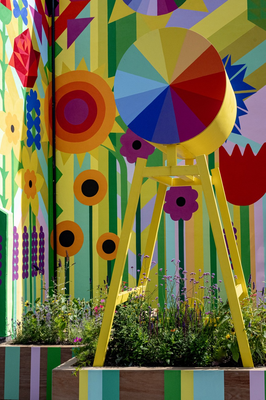 Colourful mural with large, bright flowers covers a wall. In front, a yellow tripod structure holds a circular disc painted with rainbow wedges, surrounded by flower beds with green plants and wildflowers.