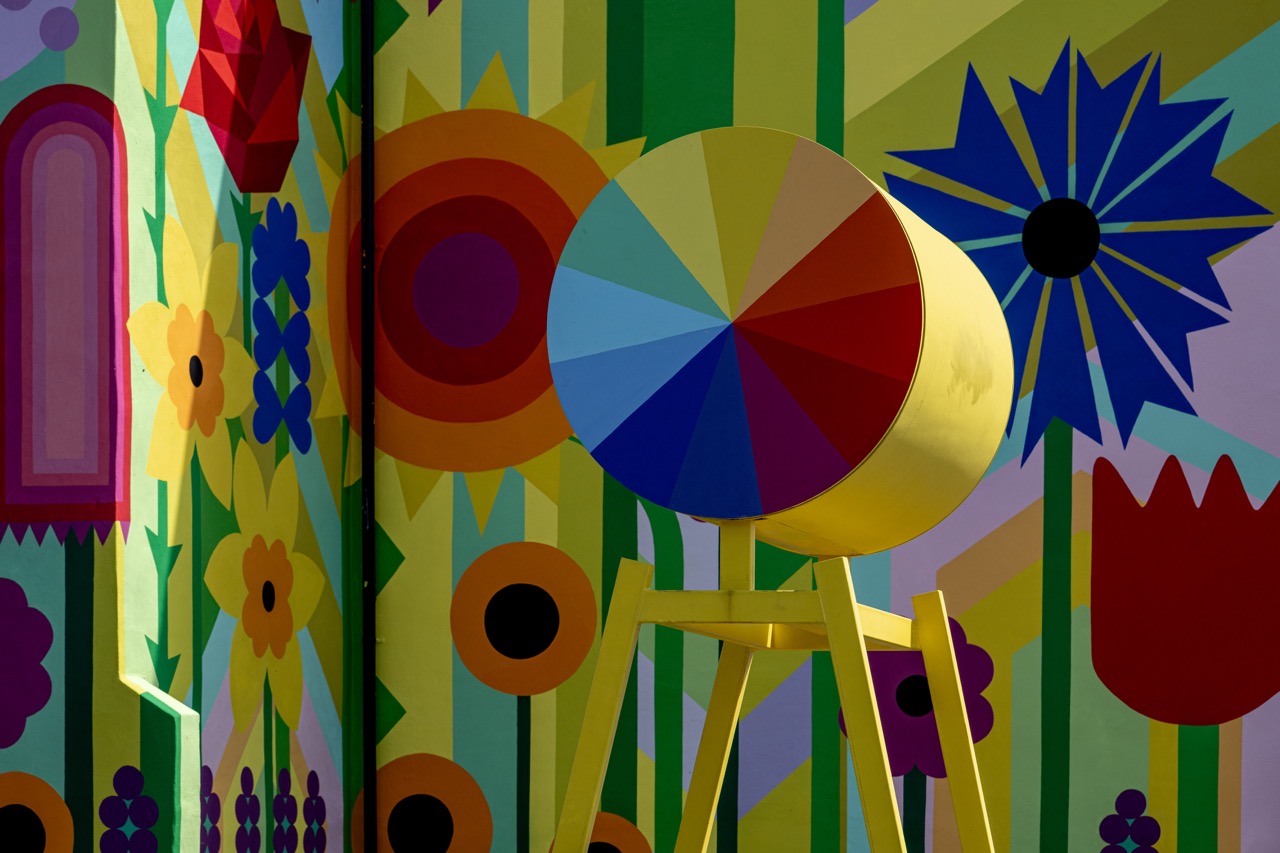 A colourful geometric sculpture with a multicoloured circular face stands against a bright mural featuring stylised flowers and bold shapes in yellow, green, purple, red, and blue.