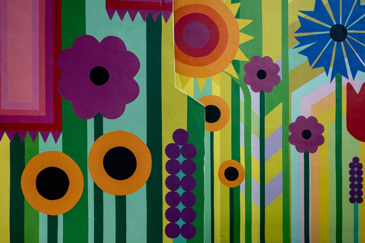 A colourful mural featuring bold, geometric flowers in shades of purple, orange, blue, red, and yellow on a background of green vertical stripes and zigzag patterns.