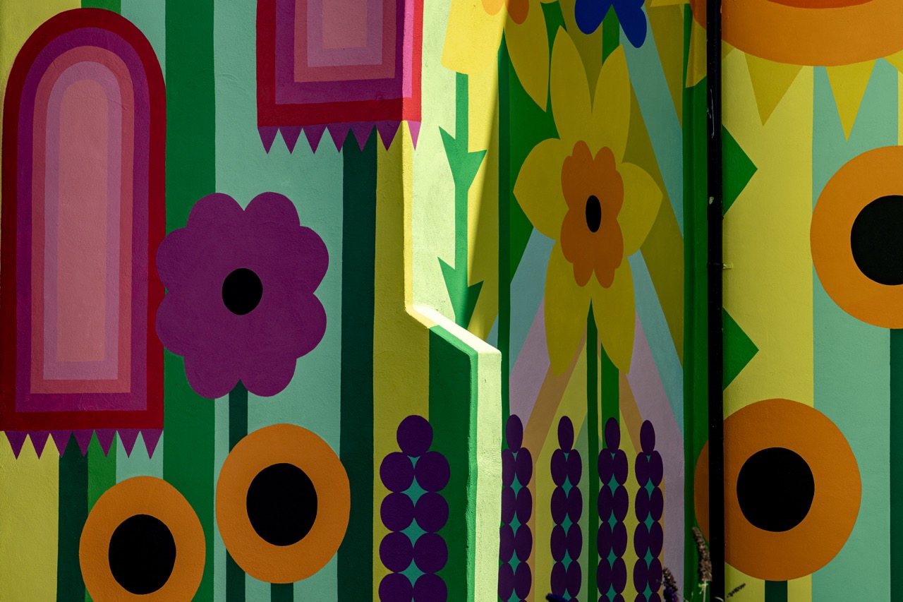 A colourful mural with bold geometric flowers in yellow, purple, black, pink, and green, featuring vertical stripes and abstract stems on a sunlit wall.