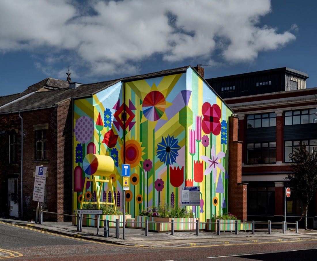 A vibrant mural with large colourful flowers, geometric shapes, and bold patterns covers the entire side of a building at a street corner under a partly cloudy sky. An art installation stands in front.