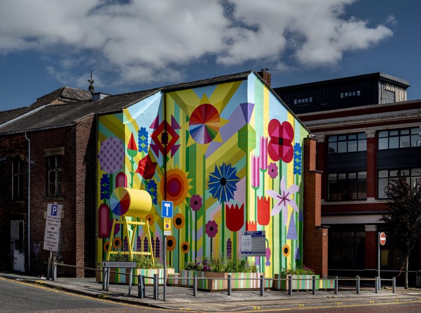 A vibrant mural with large colourful flowers, geometric shapes, and bold patterns covers the entire side of a building at a street corner under a partly cloudy sky. An art installation stands in front.
